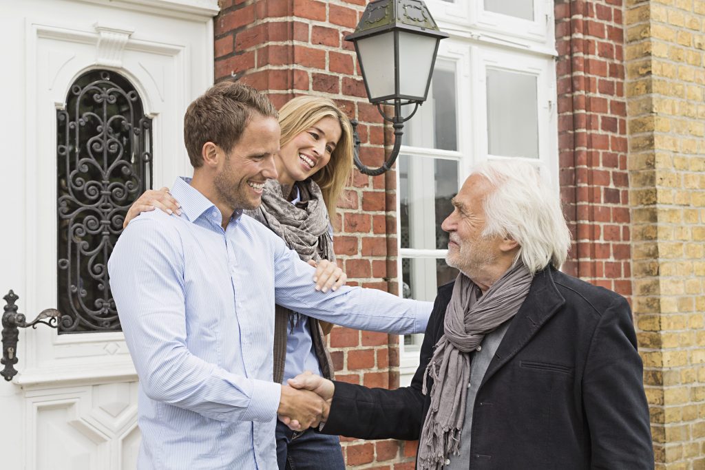 Couple souriant serrant la main d’un homme âgé devant une maison en briques