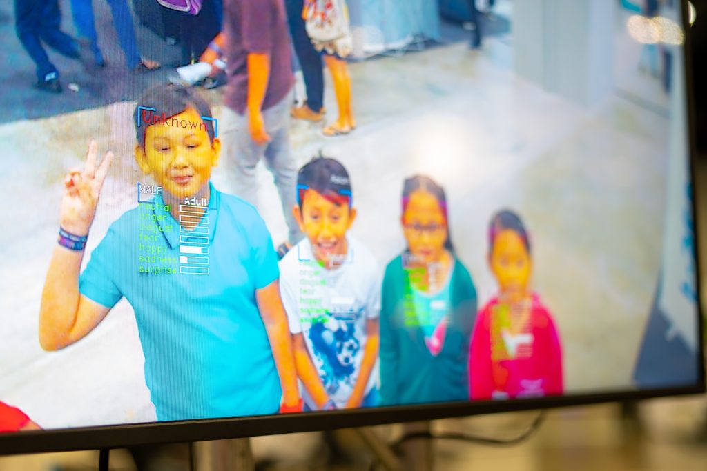 Écran affichant un logiciel de reconnaissance faciale analysant les visages et les émotions de plusieurs enfants dans un lieu public.